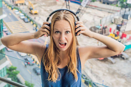A young woman by the window annoyed by the building works outside, wired soundproof wireless headphones.の写真素材