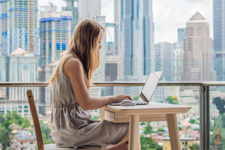 Young woman is working on a laptop on her balcony overlooking the skyscrapers. Freelancer, remote work, work from home.の写真素材