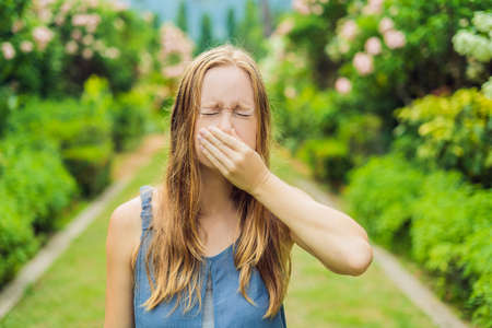 Young woman sneezes in the park against the background of a flowering tree. Allergy to pollen...