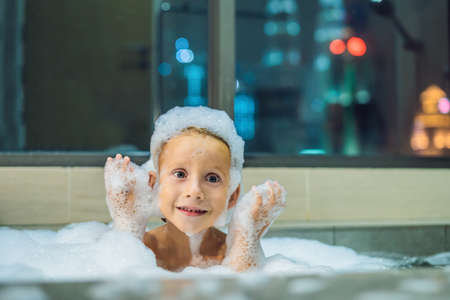 Happy little baby boy sitting in bath tub in the evening before going to sleep on the background of a window overlooking the evening city. Portrait of baby bathing in a bath full of foam near window.の写真素材