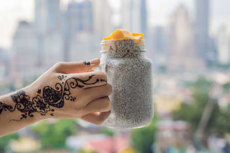 Young woman with mehendi from henna on hand eating chia pudding on her balcony overlooking the big city.の写真素材