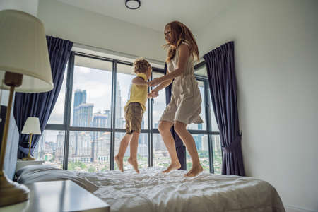 Happy mother and son jumping on the bed against the backdrop of a large window overlooking the skyscrapers and the big city.の写真素材