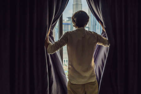 Young man opens the window curtains and looks at the skyscrapers in the big city.の写真素材