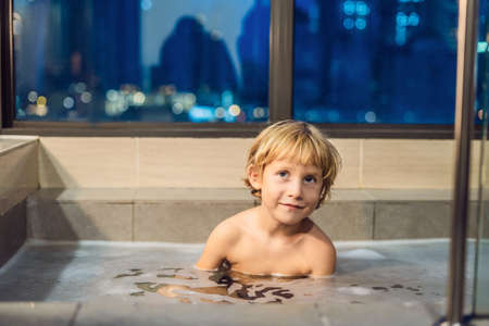 Happy little baby boy sitting in bath tub in the evening before going to sleep on the background of a window overlooking the evening city. Portrait of baby bathing in a bath full of foam near window.の写真素材