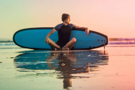 Silhouette of surf man sitting with a surfboard on the seashore beach at sunset time.