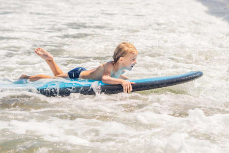 Happy baby boy - young surfer ride on surfboard with fun on sea waves. Active family lifestyle, kids outdoor water sport lessons and swimming activity in surf camp. Summer vacation with child.の写真素材
