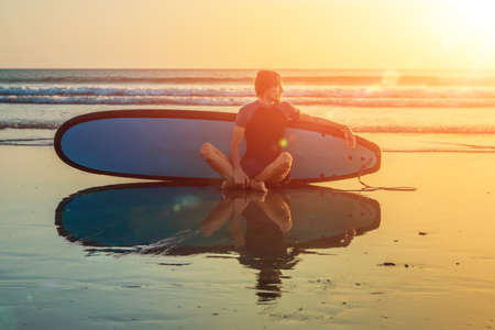 Silhouette of surf man sitting with a surfboard on the seashore beach at sunset time.の写真素材