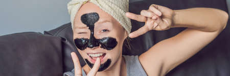 Beautiful young woman relaxing with face mask at beauty spa. Happy joyful woman applying black mask on face. BANNER long formatの写真素材