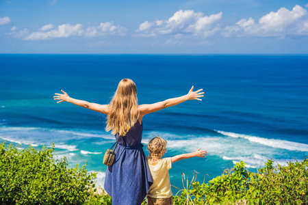 Mom and son travelers on a cliff above the beach. Empty paradise beach, blue sea waves in Bali island, Indonesia.の写真素材