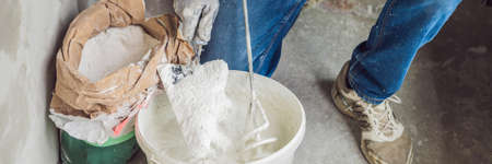 Young male painter kneads putty with water in a bucket using a hand-held mixer for building mixes. BANNER, LONG FORMATの写真素材