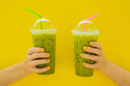 Two Green tea latte with ice in plastic cup and straw in female hand with yellow manicure on yellow background. Homemade Iced Matcha Latte Tea with Milk take awayの写真素材