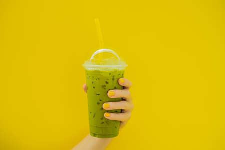 Green tea latte with ice in plastic cup and straw in female hand with yellow manicure on yellow background. Homemade Iced Matcha Latte Tea with Milk take awayの写真素材