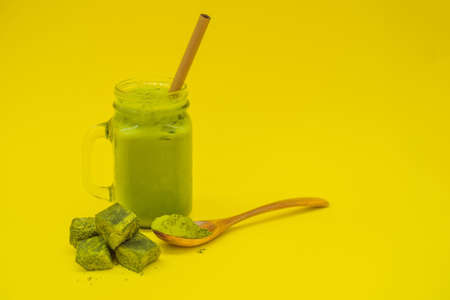 Green tea latte with ice in mason jar and straw and spoon with powder matcha and candy made of matcha on yellow background. Homemade Iced Matcha Latte Tea with Milk zero wasteの写真素材