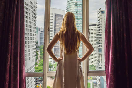 Young woman opens the window curtains and looks at the skyscrapers in the big cityの写真素材