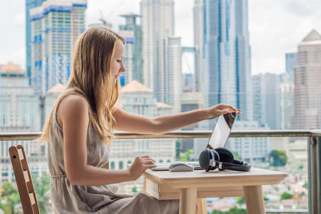 Young woman is working on a laptop on her balcony overlooking the skyscrapers. Freelancer, remote work, work from homeの写真素材