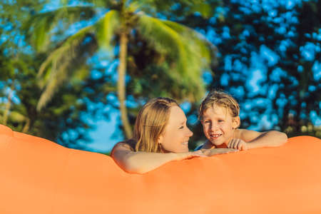 Summer lifestyle portrait of mother and son sitting on the orange inflatable sofa on the beach of tropical island. Relaxing and enjoying life on air bedの写真素材