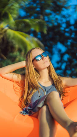 Summer lifestyle portrait of pretty girl sitting on the orange inflatable sofa on the beach of...