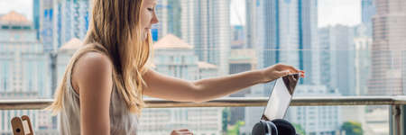 Young woman is working on a laptop on her balcony overlooking the skyscrapers. Freelancer, remote work, work from home BANNER, LONG FORMATの写真素材