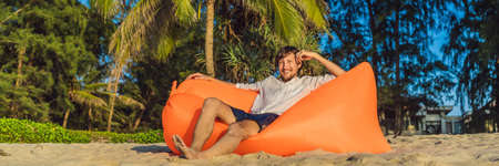 Summer lifestyle portrait of man sitting on the orange inflatable sofa on the beach of tropical island. Relaxing and enjoying life on air bed BANNER, LONG FORMATの写真素材