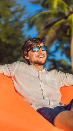 Summer lifestyle portrait of man sitting on the orange inflatable sofa on the beach of tropical island. Relaxing and enjoying life on air bed VERTICAL FORMAT for mobile story or stories size. Mobile wallpaperの写真素材