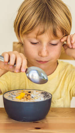 Happy boy sitting and eating a fresh smoothie bowl with mango, dragon fruit, granola and chia seeds...