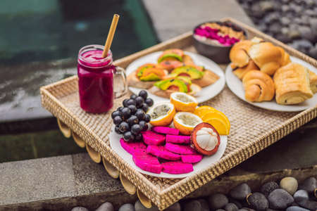 Breakfast on a tray with fruit, buns, avocado sandwiches, smoothie bowl by the pool. Summer healthy diet, vegan breakfast. Tasty vacation conceptの写真素材