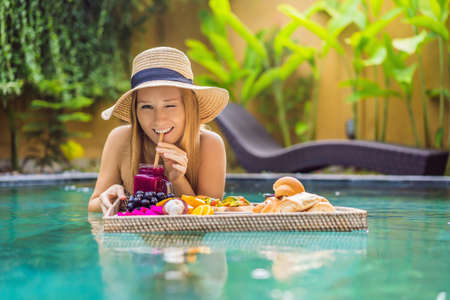 Breakfast tray in swimming pool, floating breakfast in luxury hotel. Girl relaxing in the pool drinking smoothies and eating fruit plate, smoothie bowl by the hotel pool. Exotic summer diet. Tropical beach lifestyle. Bali Trendの写真素材
