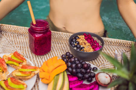 Breakfast tray in swimming pool, floating breakfast in luxury hotel. Girl relaxing in the pool drinking smoothies and eating fruit plate, smoothie bowl by the hotel pool. Exotic summer diet. Tropical beach lifestyle. Bali Trendの写真素材