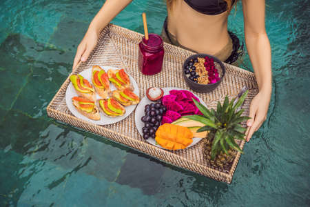 Breakfast tray in swimming pool, floating breakfast in luxury hotel. Girl relaxing in the pool drinking smoothies and eating fruit plate, smoothie bowl by the hotel pool. Exotic summer diet. Tropical beach lifestyle. Bali Trendの写真素材