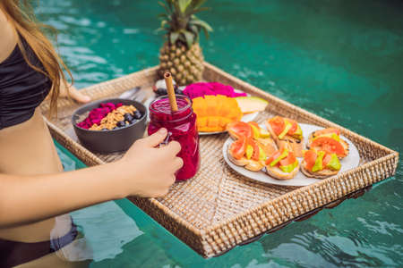 Breakfast tray in swimming pool, floating breakfast in luxury hotel. Girl relaxing in the pool drinking smoothies and eating fruit plate, smoothie bowl by the hotel pool. Exotic summer diet. Tropical beach lifestyle. Bali Trendの写真素材