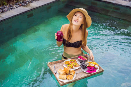Breakfast tray in swimming pool, floating breakfast in luxury hotel. Girl relaxing in the pool drinking smoothies and eating fruit plate, smoothie bowl by the hotel pool. Exotic summer diet. Tropical beach lifestyle. Bali Trendの写真素材