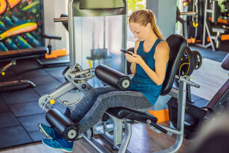 Young woman using phone while training at the gym. Woman sitting on exercising machine holding...