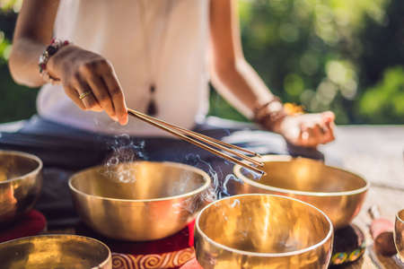 Woman playing on Tibetan singing bowl while sitting on yoga mat against a waterfall. Vintage tonned. Beautiful girl with mala beads meditatingの写真素材