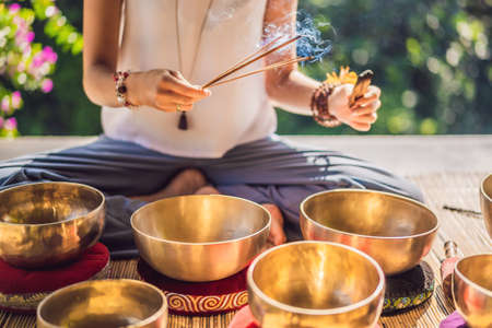 Woman playing on Tibetan singing bowl while sitting on yoga mat against a waterfall. Vintage tonned. Beautiful girl with mala beads meditatingの写真素材