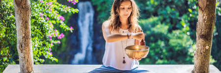 Woman playing on Tibetan singing bowl while sitting on yoga mat against a waterfall. Vintage tonned. Beautiful girl with mala beads meditating BANNER, LONG FORMATの写真素材