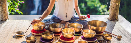 Woman playing on Tibetan singing bowl while sitting on yoga mat against a waterfall. Vintage tonned. Beautiful girl with mala beads meditating BANNER, LONG FORMATの写真素材