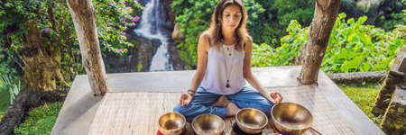 Woman playing on Tibetan singing bowl while sitting on yoga mat against a waterfall. Vintage tonned. Beautiful girl with mala beads meditating BANNER, LONG FORMATの写真素材