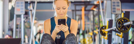 Young woman using phone while training at the gym. Woman sitting on exercising machine holding...
