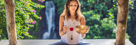 Woman playing on Tibetan singing bowl while sitting on yoga mat against a waterfall. Vintage tonned. Beautiful girl with mala beads meditating BANNER, LONG FORMATの写真素材