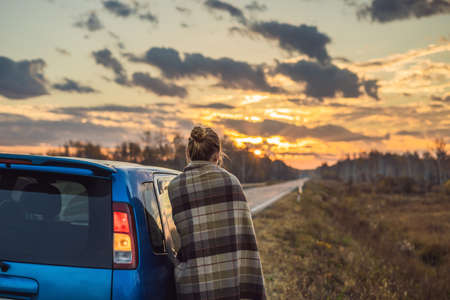 A woman in a plaid stands by the car on the side of the road in the background of the dawn. Road trip concept.の写真素材