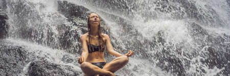 Young woman sitting in lotus position on the rock in tropical waterfallの写真素材