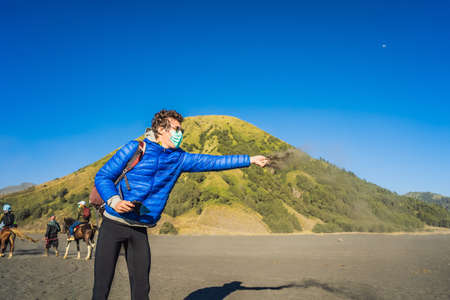 Young man tourist touches volcanic sand in the Bromo Tengger Semeru National Park on the Java Island, Indonesia. He enjoys magnificent view on the Bromo or Gunung Bromo on Indonesian, Semeru and other volcanoes located inside of the Sea of Sand within the Tengger Caldera. One of the most famous volcanic objects in the world. Travel to Indonesia conceptの写真素材