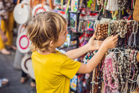Boy at a market in Ubud, Bali. Typical souvenir shop selling souvenirs and handicrafts of Bali at the famous Ubud Market, Indonesia. Balinese market. Souvenirs of wood and crafts of local residents. Traveling with children concept. Kids friendly placeの写真素材