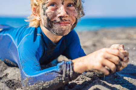 Black Friday concept. Smiling boy with dirty Black face sitting and playing on black sand sea beach before swimming in ocean. Family active lifestyle, and water leisure on summer vacation with kids. Black Friday, sales of tours and airline tickets or goodsの写真素材