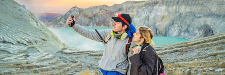BANNER, LONG FORMAT Young tourist man and woman stand at the edge of the crater of the Ijen volcano or Kawah Ijen on the Indonesian language. Famous volcano containing the biggest in the world acid lake and sulfur mining spot at the place where volcanic gasses come from the volcano. They wear respirators to protect from dangerous volcanic gassesの写真素材