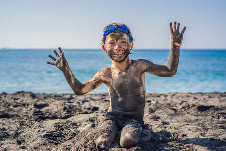 Black Friday concept. Smiling boy with dirty Black face sitting and playing on black sand sea beach before swimming in ocean. Family active lifestyle, and water leisure on summer vacation with kids. Black Friday, sales of tours and airline tickets or goodsの写真素材