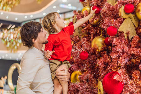Father and son hangs a decoration on the red Christmas tree. Red Christmas tree on the background of lightsの写真素材