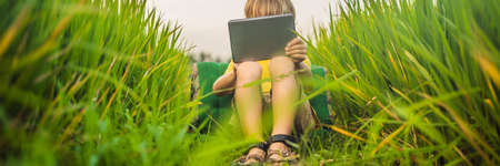 Happy child sitting on the field holding tablet. Boy sitting on the grass on sunny day. Home schooling or playing a tablet BANNER, LONG FORMATの写真素材