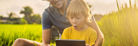 Dad and son sitting on the field holding tablet. Boy sitting on the grass on sunny day. Home...