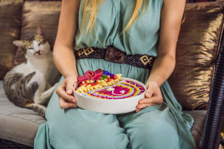 Young woman having a mediterranean breakfast seated at her sofa and eats Healthy tropical breakfast, smoothie bowl with tropical fruits, decorated with a pattern of colorful yogurt with turmeric and spirulina. It is also decorated with fruits, flowers, chia seeds, coconut, granola, pineapple, mint and strawberriesの写真素材